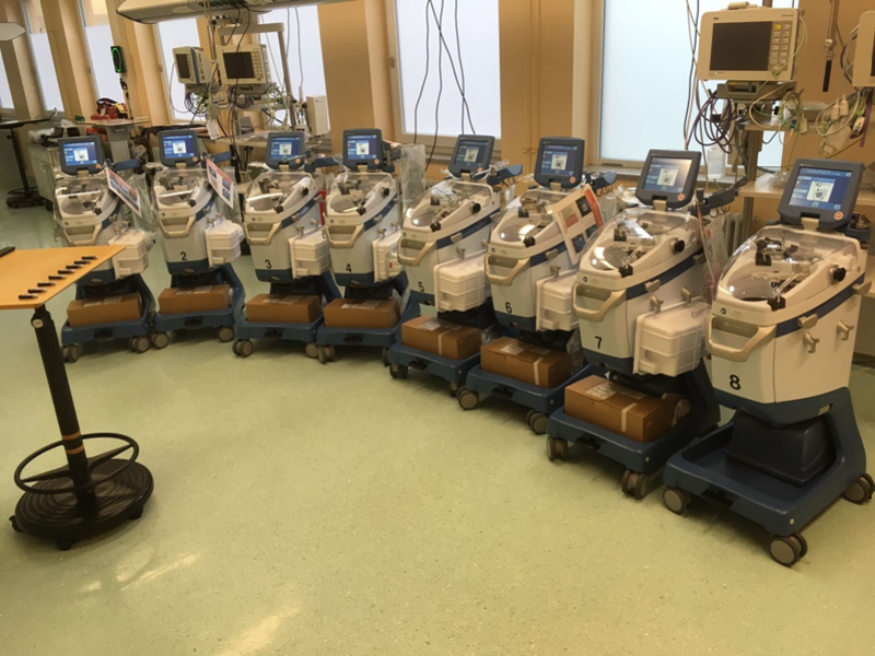 Reihe von acht medizinischen Geräten zur maschinellen Blutverarbeitung mit Monitoren, aufgereiht in einem Klinikraum.