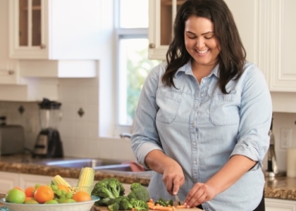 Junge Frau schneidet in einer Küche auf einem Holzbrett Gemüse. Eine Schale mit verschiedenen Obst- und Gemüsesorten steht auf der Arbeitsplatte.